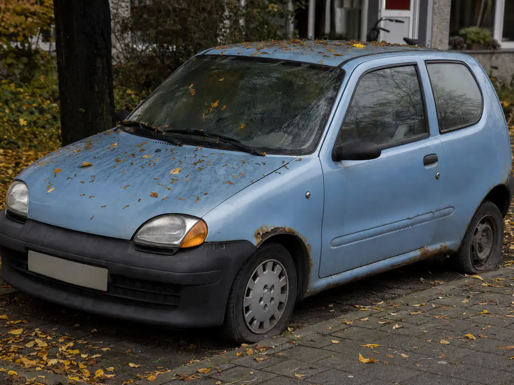 Oude lichtblauwe hatchback met roest en een zachte achterband, lang geparkeerd onder herfstbladeren